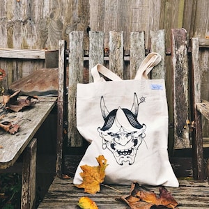 May include: A white tote bag featuring a black and white illustration of a Japanese demon mask. The bag has a blue tag with the text "Artist's Touch" on it.