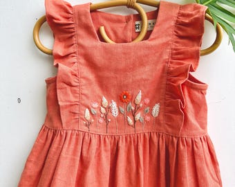 May include: Coral-colored dress with floral embroidery. The dress features a square neckline, ruffled sleeves, and a gathered skirt. The embroidery includes a central orange flower and various other flowers and leaves.