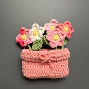 May include: A handmade crochet flower pot with colorful flowers. The pot is a soft pink color with a bow detail. The flowers are pink, white, and red with yellow centers and green leaves. The flowers are arranged in a decorative display.