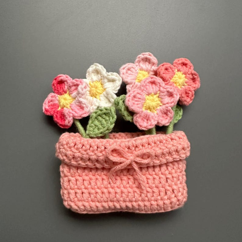May include: A handmade crochet flower pot with colorful flowers. The pot is a soft pink color with a bow detail. The flowers are pink, white, and red with yellow centers and green leaves. The flowers are arranged in a decorative display.
