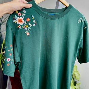 May include: A dark green t-shirt with embroidered flowers in shades of pink, yellow, and white on the left shoulder.