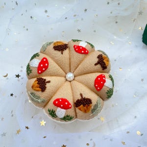 May include: A beige fabric pincushion with a pearl button in the center. The pincushion is decorated with embroidered mushrooms and acorns.