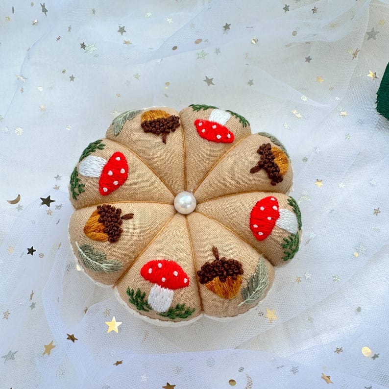 May include: A beige fabric pincushion with a pearl button in the center. The pincushion is decorated with embroidered mushrooms and acorns.