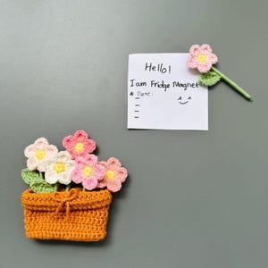 May include: A handmade crochet flower pot magnet with pink, white, and yellow flowers. The orange pot has a tied bow. A separate flower magnet is attached to a note that says "Hello! I am Fridge Magnet."