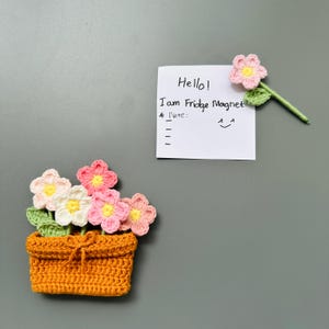 May include: A crocheted orange flower pot with pink, white, and yellow flowers. A single crocheted pink flower with a green stem and leaf is also present. A white note says "Hello! I am Fridge Magnet."