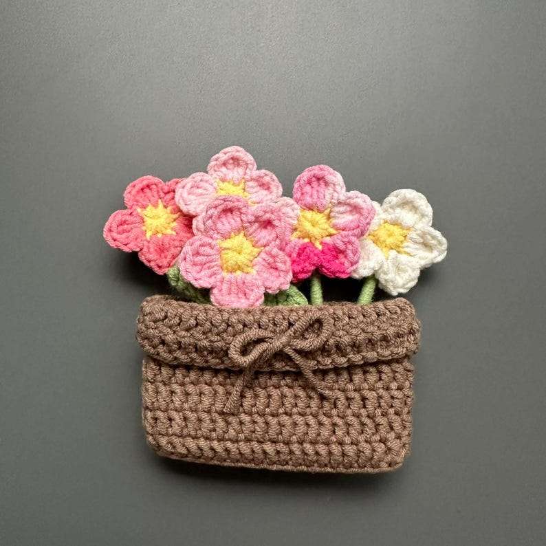 May include: A handmade crochet flower arrangement in a brown basket. The flowers are pink, white, and red with yellow centers. The basket has a decorative bow. The craft is set against a gray background.