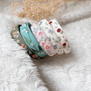 May include: Five embroidered headbands in different floral patterns. The headbands are stacked on top of each other, with the green headband at the bottom and the white headband at the top. The headbands are all made of a soft, white fabric and feature delicate floral embroidery.