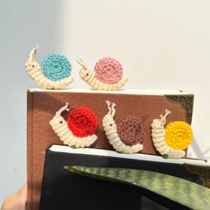 May include: Five crocheted snail bookmarks in different colors. The snails are white with colored shells. The shells are blue, pink, red, brown, and yellow.