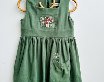 May include: A green sleeveless dress with a square neckline and a gathered skirt. The dress features an embroidered mushroom and floral design on the bodice and a pocket with a floral design. The dress is hanging on a wooden hanger.