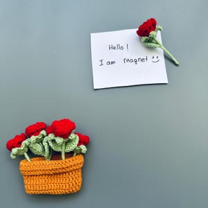 May include: Crocheted magnets featuring a red rose and a pot of red roses. The pot is orange and the leaves are green. A white note says "Hello! I am Magnet".