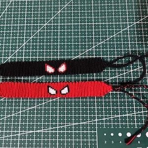 Può includere: Due braccialetti macramè fatti a mano. Uno è nero con dettagli a forma di occhi bianchi e rossi, l'altro è rosso con dettagli a forma di occhi bianchi e neri. Entrambi hanno lacci regolabili e sono esposti su un tappetino da taglio verde.
