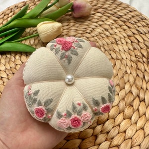 May include: A white fabric pincushion with a pearl button in the center and embroidered pink flowers and green leaves.
