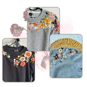 May include: Three sweatshirts with embroidered floral designs. The first sweatshirt is gray with yellow, orange, and white flowers. The second sweatshirt is black with pink and white flowers. The third sweatshirt is blue with pink, white, and green flowers.