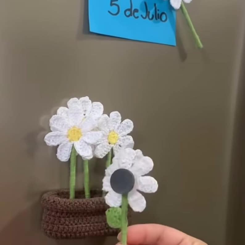 May include: Crocheted daisy magnets with white petals and yellow centers. One is attached to a brown crocheted pot, while another is held up. A blue sticky note with the text "5 de Julio" is visible.