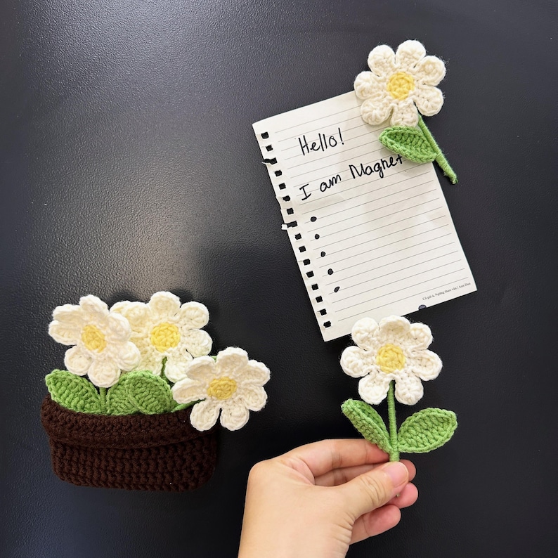 May include: Crocheted daisy magnets with white petals and yellow centers, along with a brown crocheted flower pot. A piece of paper with the text "Hello! I am Magnet" is also present.
