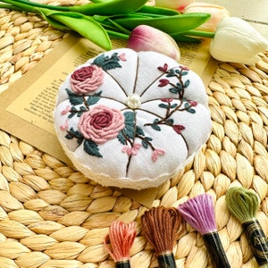 May include: White fabric pincushion with a lace trim, embroidered with pink and green flowers. The pincushion is sitting on a woven wicker surface with four spools of embroidery floss in pink, brown, green, and purple.