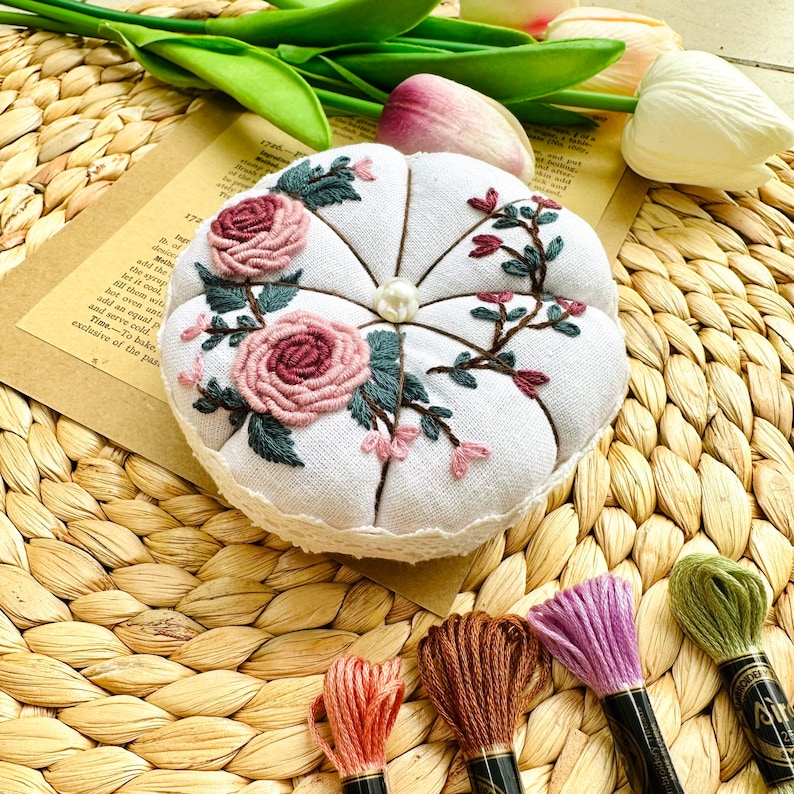 May include: White fabric pincushion with a lace trim, embroidered with pink and green flowers. The pincushion is sitting on a woven wicker surface with four spools of embroidery floss in pink, brown, green, and purple.