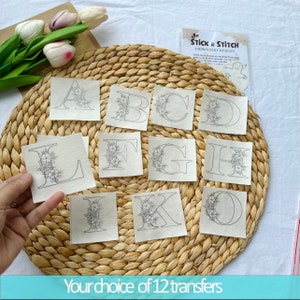 Può includere: Dodici fogli di trasferimento per ricamo con lettere floreali dalla A alla L. Ogni foglio è un quadrato con sfondo bianco e contorno nero di una lettera con accenti floreali. I fogli sono disposti su un tappetino di paglia intrecciata.