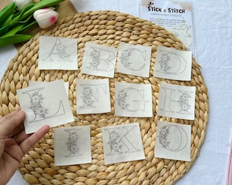 May include: Twelve embroidery transfer sheets featuring floral letters A through L. Each sheet is a square with a white background and black outline of a letter with floral accents. The sheets are arranged on a woven straw mat.