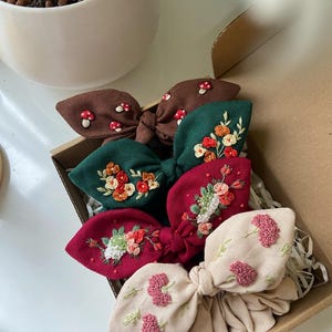 May include: Set of four handmade hair scrunchies with embroidered floral and mushroom designs. The scrunchies are in shades of brown, green, red, and beige.