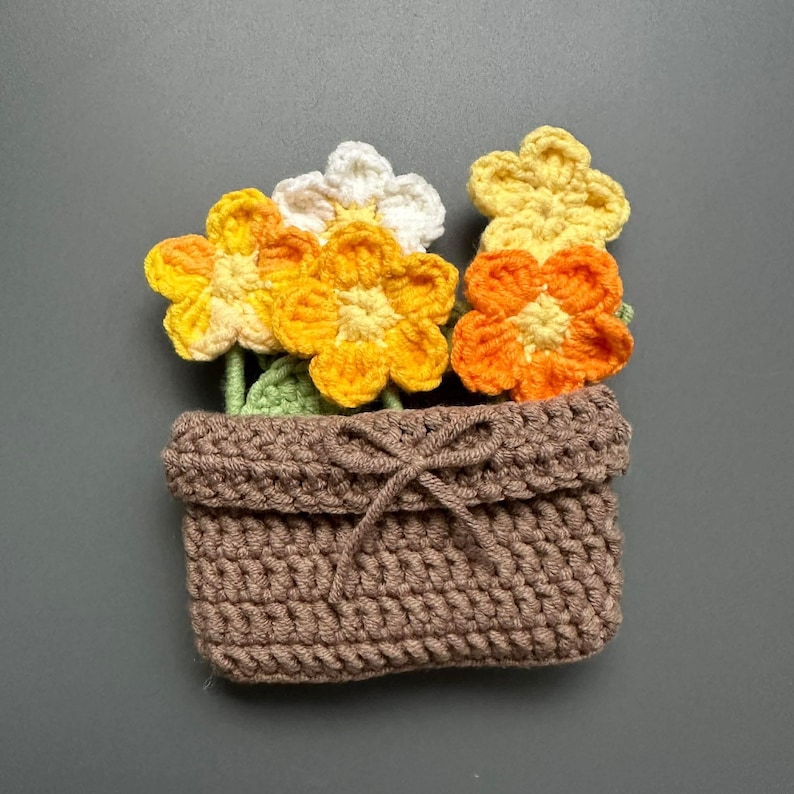May include: A crocheted flower pot with a bow, filled with colorful crocheted flowers. The flowers are yellow, orange, and white, with green leaves. The pot is brown.