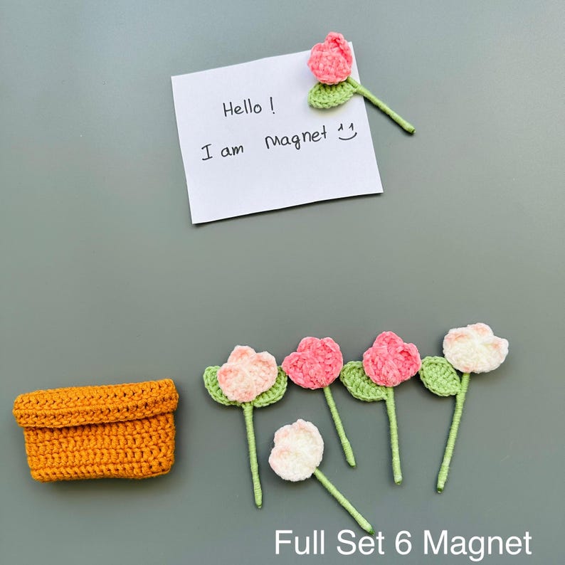 May include: A set of six handmade crochet flower magnets in shades of pink and white, with green leaves and stems. Includes a small orange crochet basket and a note that says "Hello! I am Magnet".