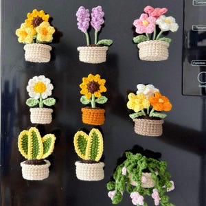 May include: A collection of handmade crochet flower magnets in various styles. The magnets feature sunflowers, daisies, and other flowers in small woven baskets. The color palette includes yellow, pink, white, and green, with brown and beige accents.