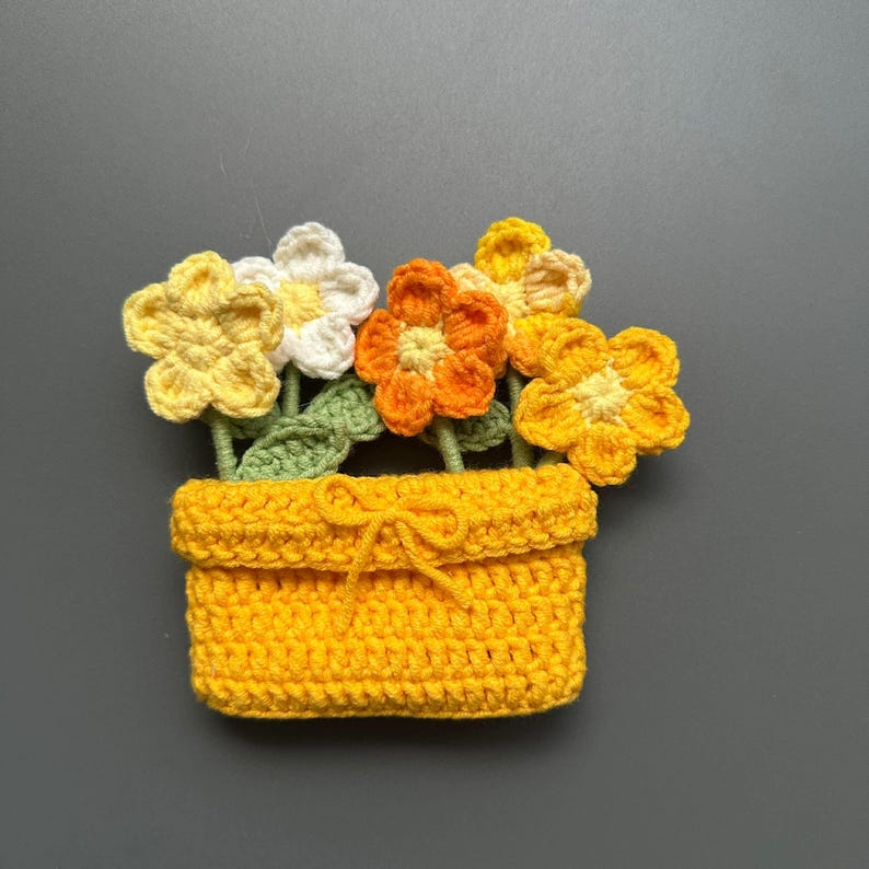 May include: A handmade crochet flower pot with yellow, orange, and white flowers. The flowers have green stems and leaves. The pot is yellow and has a bow. The craft is set against a gray background.