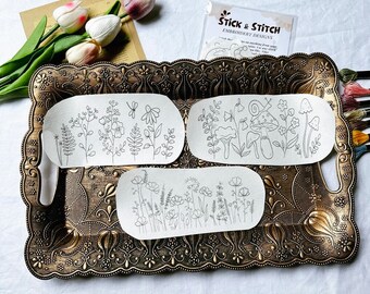 May include: Three embroidery patterns on white paper featuring floral and mushroom designs. The patterns are on a gold tray with a decorative border. The text on the paper reads "Stick & Stitch Embroidery Designs".