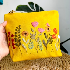 May include: A yellow canvas pouch with embroidered flowers in shades of pink, orange, yellow, and green. The pouch has a zipper closure.