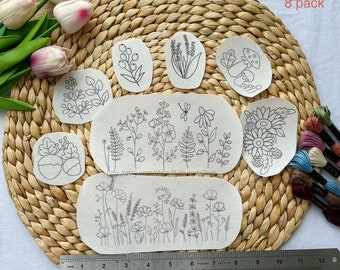 May include: Eight pack of embroidery patterns featuring various floral designs, including a sunflower, mushrooms, and a variety of wildflowers. The patterns are printed on white fabric and are ready to be embroidered.