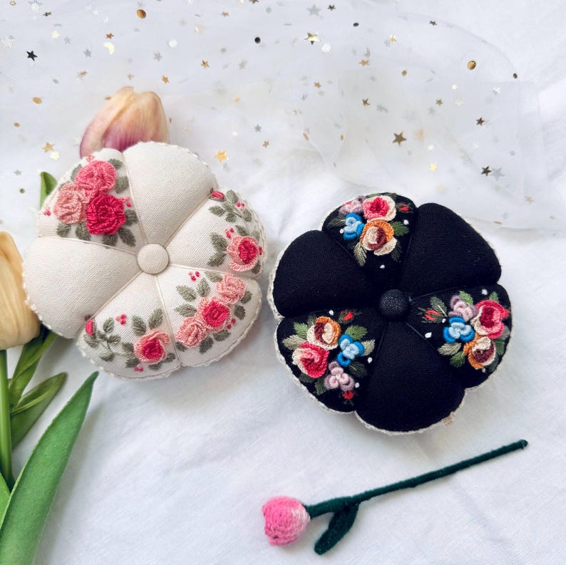 May include: Two floral embroidered pincushions, one white and one black, with a small pink flower on a stick.