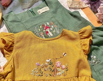 May include: Two children's dresses. The front dress is mustard yellow with embroidered flowers and a bee. The second dress is green with embroidered mushrooms. Both dresses have a square neckline and ruffled sleeves. The dresses are made of linen.