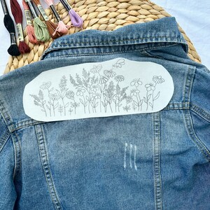 May include: A denim jacket with a white fabric patch on the back. The patch has a line drawing of a floral design with a butterfly.
