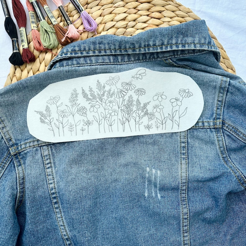 May include: A denim jacket with a white fabric patch on the back. The patch has a line drawing of a floral design with a butterfly.