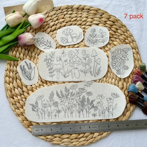 May include: Seven pack of embroidery patterns featuring floral and mushroom designs. The patterns are printed on white fabric and are ready to be stitched. The patterns are arranged on a woven straw mat.