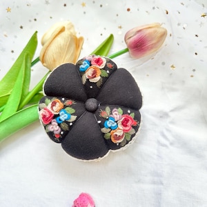 May include: A black fabric pincushion with a white lace trim and embroidered flowers in shades of pink, blue, and yellow.