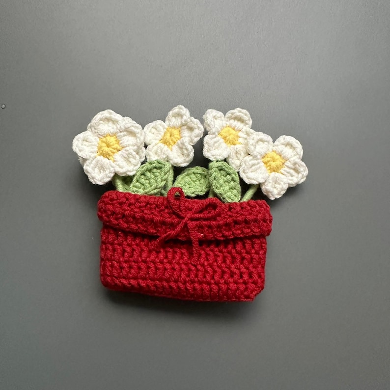 May include: A handmade crochet flower pot with white flowers, yellow centers, and green leaves. The pot is red and tied with a red bow. The flowers are arranged in a decorative pattern.