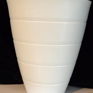 以下が含まれることがあります： 水平の溝が入った白い先細りの花瓶。花瓶は広い開口部とわずかにテクスチャ加工された表面を持っています。 デザインはシンプルでモダンで、さまざまな家の装飾スタイルに適しています。