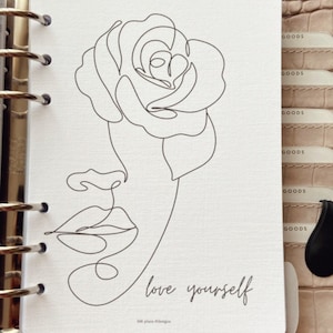 Pode incluir: Uma inserção de planejador branca com um desenho de linha preta de uma rosa e um rosto. As palavras "love yourself" estão escritas em uma fonte de script abaixo do desenho. A inserção está em um planejador de couro marrom.