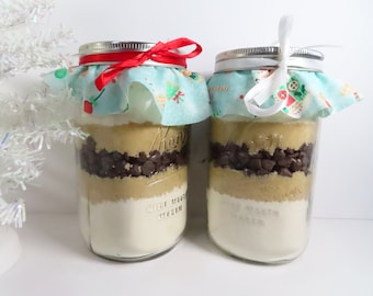May include: Two glass jars filled with cookie ingredients, layered with flour, brown sugar, chocolate chips, and a festive fabric tied around the top with a ribbon.