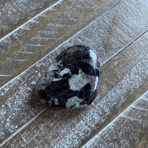 May include: A polished stone with a mix of black, white, and burgundy colors. The stone has a smooth, rounded shape and a glossy finish. The stone is likely a decorative item or a collectible.