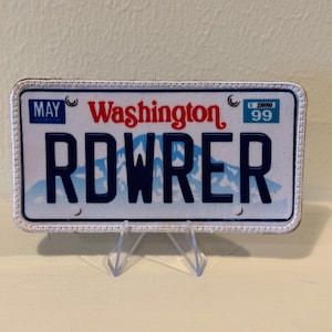 May include: A rectangular license plate with the word "Washington" in red script. The plate reads "RDWRER" in dark blue block letters over a mountain scene. The month "MAY" is in a blue box in the upper left corner. The plate is displayed on a clear stand.
