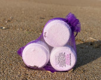May include: Three round, light pink shower steamers in a purple mesh bag, resting on a sandy beach. The steamers have a label with text, including "Shower Steamer" and "Lavender Essential Oil". The ocean is visible in the background.