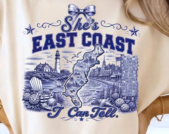 May include: A cream-colored t-shirt featuring a blue graphic print with a bow at the top, the text "She's EAST COAST I Can Tell.", a map of the East Coast, a lighthouse, coastal scenery, and a signpost listing states from Maine to Florida.