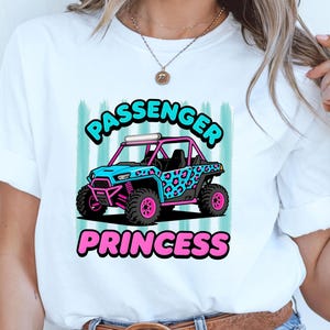 May include: A white short-sleeved t-shirt featuring a graphic of a blue and pink leopard print off-road vehicle. The text PASSENGER PRINCESS is printed in bold blue and pink letters above and below the vehicle illustration on a striped background.