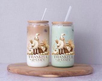 May include: Two glass tumblers with bamboo lids and clear straws, each filled with a different colored beverage. The glasses feature a graphic of a goose wearing a scarf and boots next to a basket of pumpkins, with the text THANKFUL & COZY.