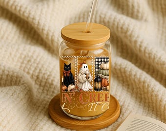 May include: A clear glass tumbler with a bamboo lid and glass straw, filled with a tan liquid. The exterior features three rectangular panels showing a black cat, a ghost holding a coffee cup, and pumpkins, with the text IN CREEP IT C.
