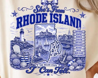 May include: A cream-colored t-shirt featuring a blue graphic print of Rhode Island. The design includes a map, a lighthouse, sailboats, a beach chair, and a list of cities including Providence, Newport, and Warwick, with the text She's From RHODE ISLAND THE OCEAN STATE I Can Tell.