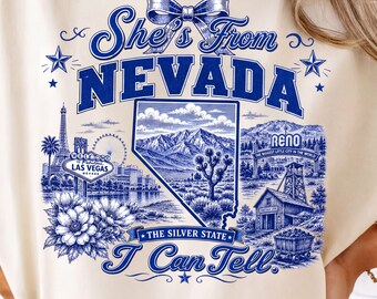 May include: A cream-colored t-shirt featuring a blue graphic print with the text She's From NEVADA, The Silver State, and I Can Tell. The design includes a bow, stars, the Las Vegas sign, the Eiffel Tower, a desert landscape, and Reno signage.